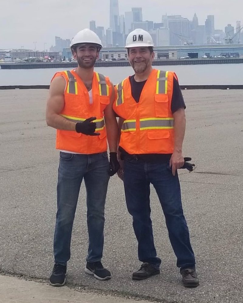 The other two family members who run D&M, in hard hats and safety vests on the Brooklyn waterfront, with Lower Manhattan behind