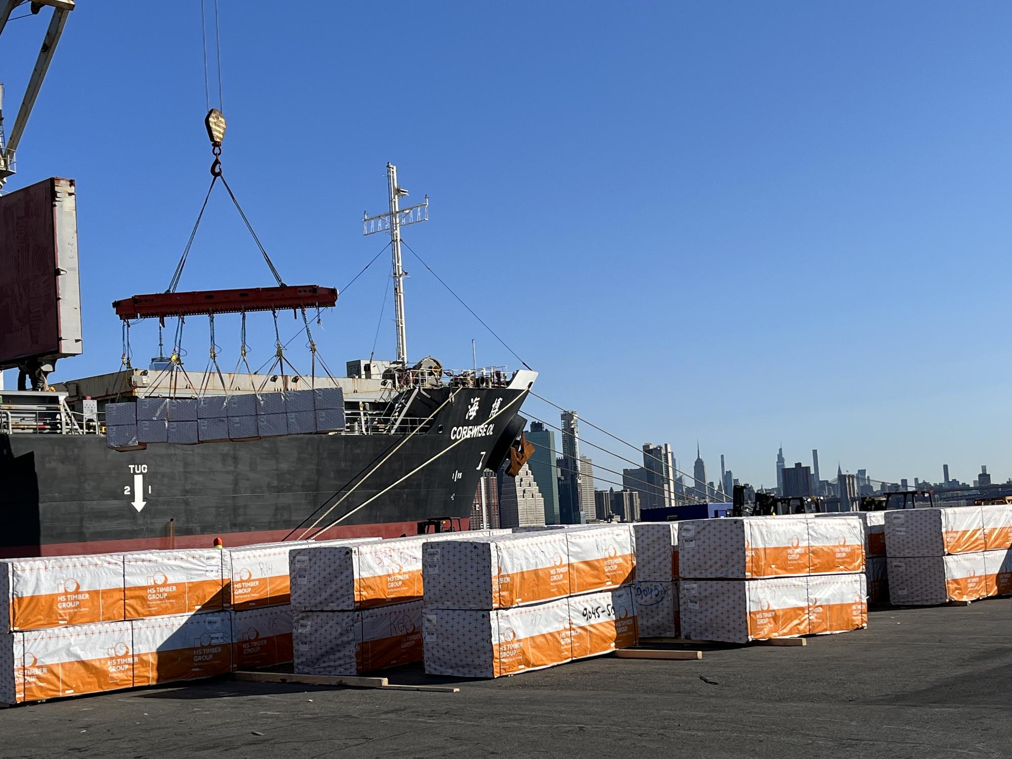 A vessel's crane lowering a set of lumber units onto the pier, wrapped units staged in rows, Manhattan skyline behind